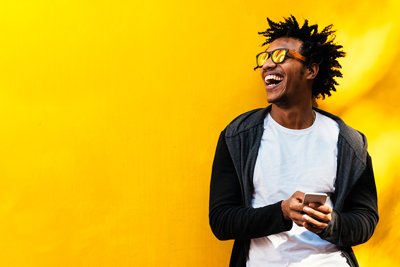 Hombre negro con gafas de sol riendo frente a una pared amarilla, con un teléfono celular en la mano