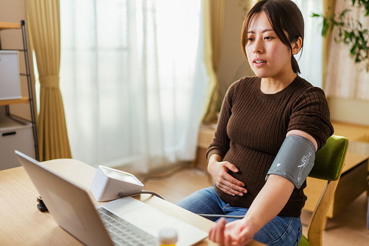 A young pregnant woman is talking with a doctor on a laptop and checking her blood pressure at home.