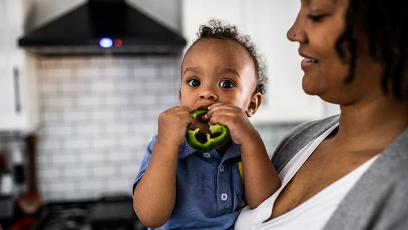 Mom holding toddler that is eating a green pepper