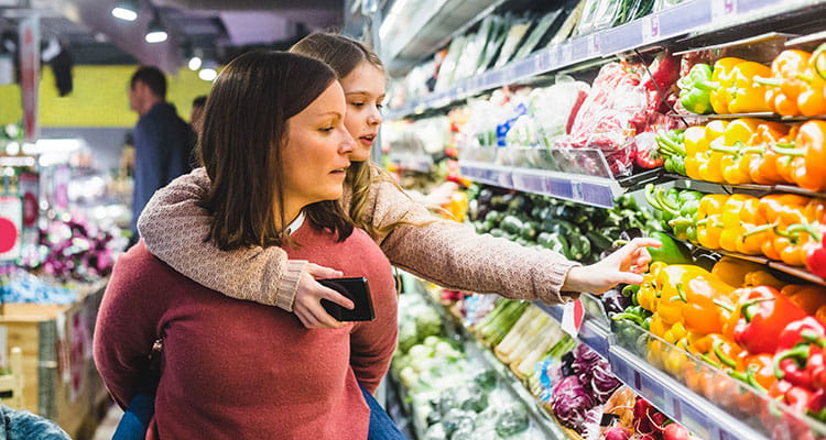 Niña eligiendo pimientos mientras su madre la lleva a caballito en el supermercado.