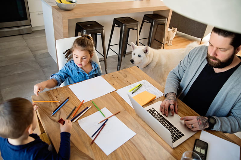 un padre utiliza su computadora portátil en la cocina mientras los niños dibujan al perro