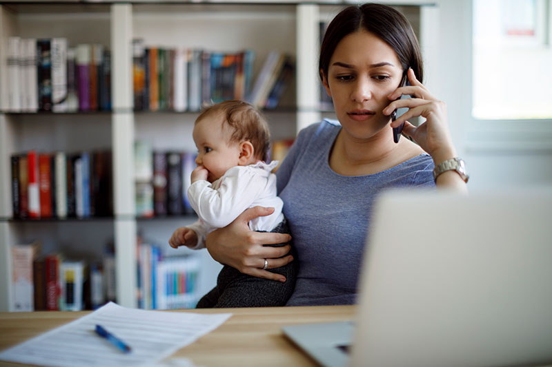 Una mamá ocupada trabajando con un teléfono celular y una computadora portátil que sostiene a su bebé
