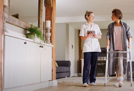 A healthcare worker and a woman with a walker are walking while chatting in a home setting.