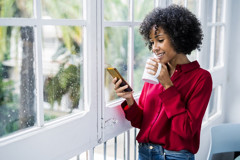 African American woman drinking coffee, looking at cell phone in front of window