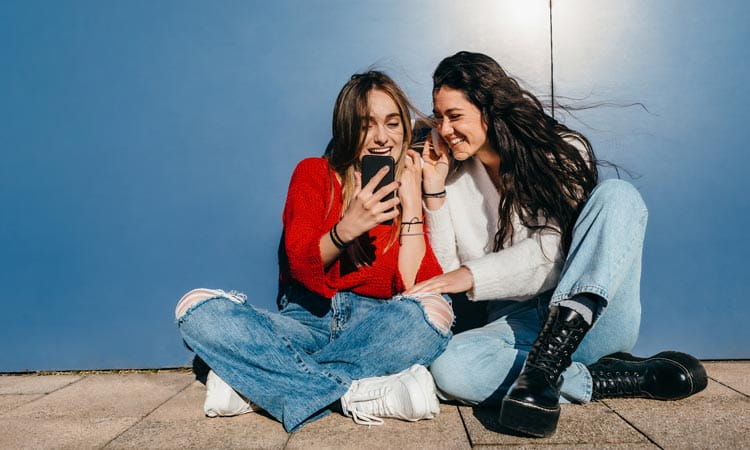 Two young women sitting on the ground outside looking at mobile device