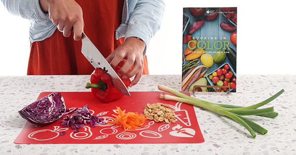 Primer plano de unas manos cortando verduras en la tabla de cortar con el libro de cocina de la American Heart Association Cooking a color